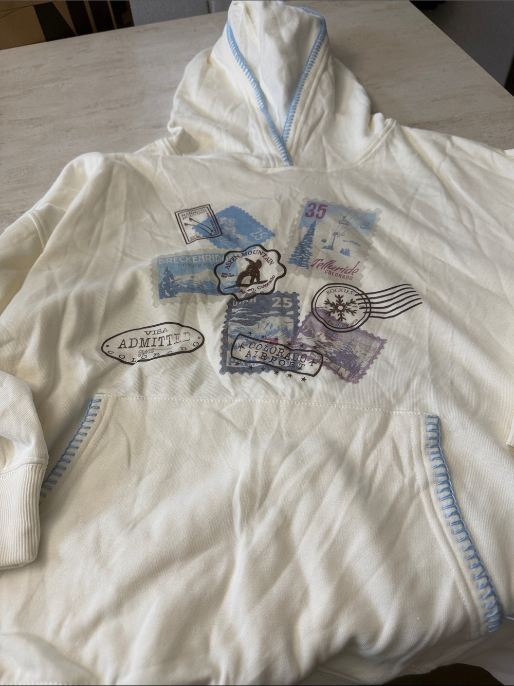 Cream Hooded Sweatshirt with Blue Trim and Travel Stamp Graphic